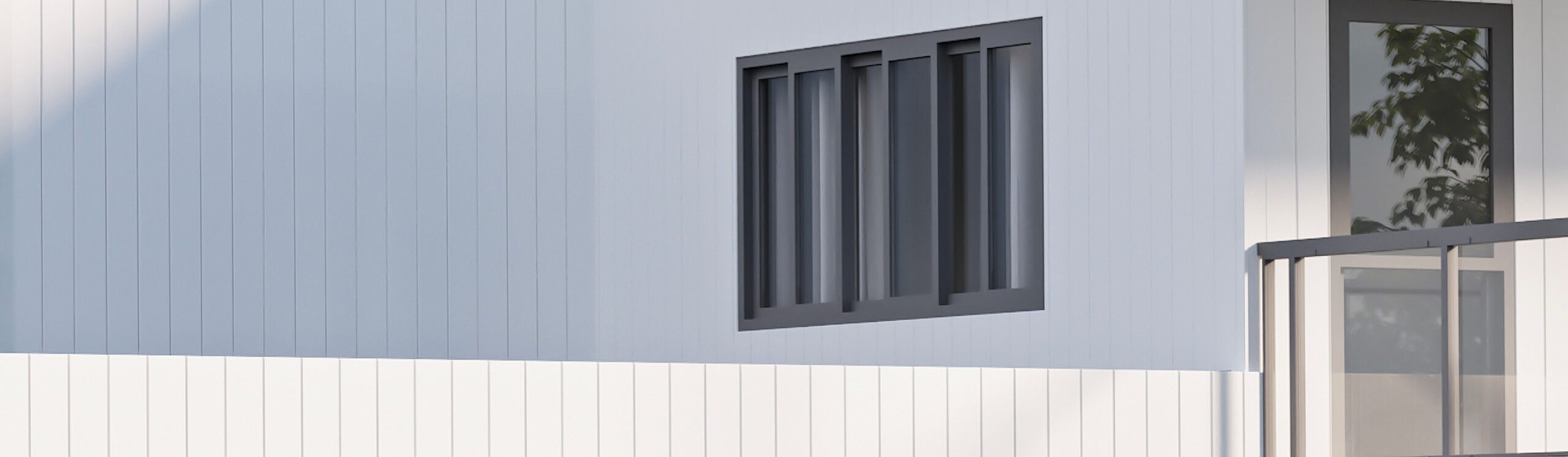 A close-up of the exterior of a modern building featuring vertical paneling in light grey and white tones. There is a set of rectangular windows with dark frames in the center. The corner of a balcony with a glass railing is visible on the right side.