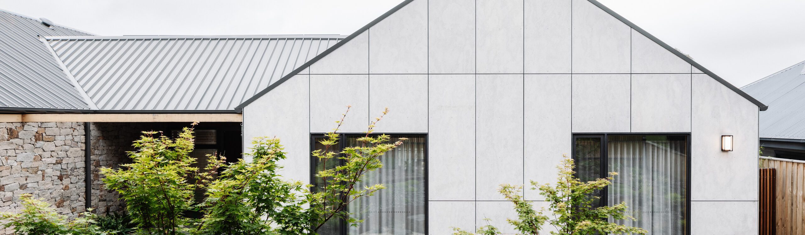 Modern house with a simple, clean design. The exterior features a combination of large, light gray panels and stone walls. The roof is corrugated metal. Several small trees with green leaves are planted in front of the house, enhancing its natural feel.