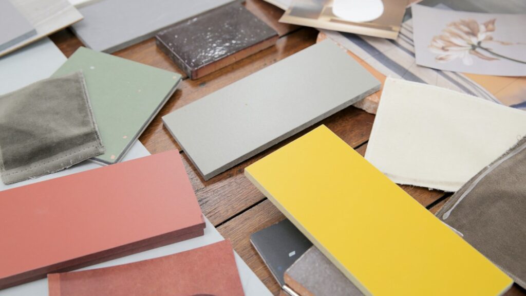 A collection of various colored and textured material samples laid out on a wooden surface. The samples include fabric swatches and rectangular pieces of different colors such as yellow, pink, green, and gray, along with some patterned samples.