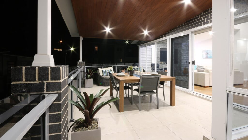 A modern, outdoor balcony at night with a wooden ceiling and recessed lights. The space features a dining table set for four, potted plants, and glass sliding doors leading to a well-lit indoor living area. The balcony has a railing with brick and metal details.