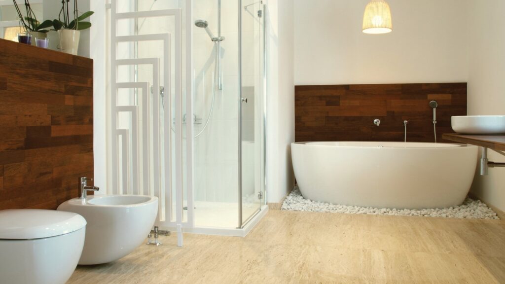 Modern bathroom with a freestanding white bathtub surrounded by decorative white pebbles, adjacent to a wooden accent wall. Features a glass-enclosed shower, white toilet, bidet, and a round vessel sink on a wooden countertop. Light fixture hangs from ceiling.