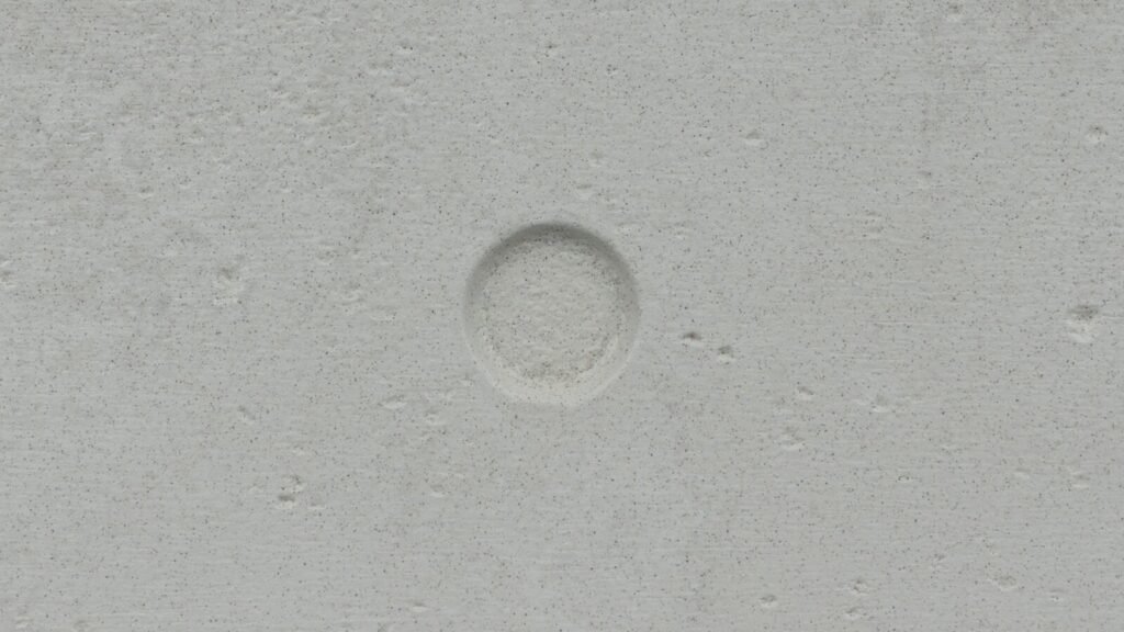 A smooth, light gray concrete surface with a shallow, circular indentation in the center. The texture is slightly rough, with small pockmarks scattered across the surface.