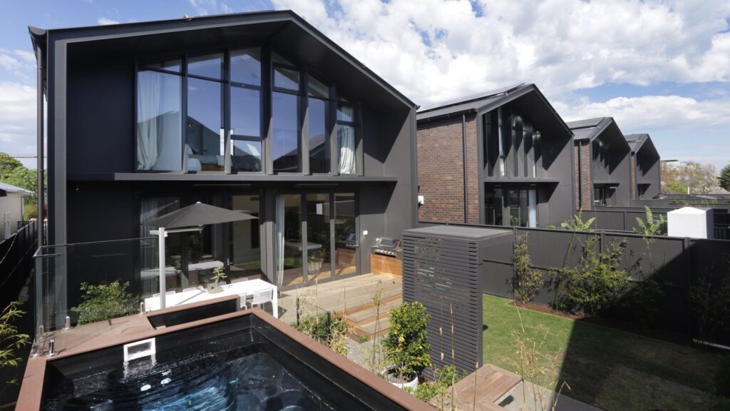 A modern backyard featuring a row of contemporary two-story homes with large glass windows, black exteriors, and wooden accents. The yard includes a small pool, wooden deck with outdoor seating and an umbrella, landscaped garden, and a glass fence.