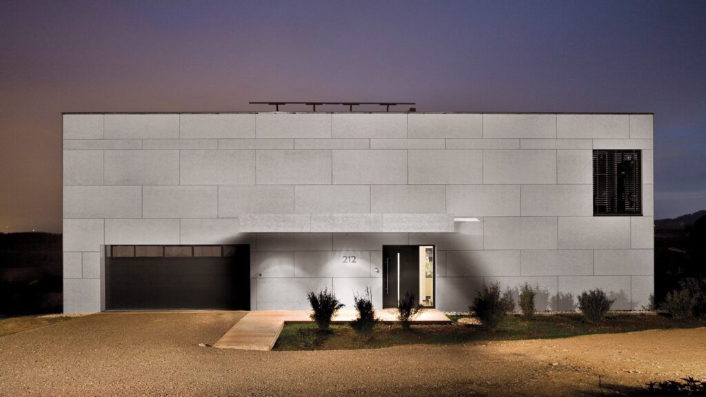 A modern, minimalist house with a flat roof, illuminated at night. The facade is composed of large, light grey, rectangular panels. The house features a recessed entrance with the number "212" on the wall, a black garage on the left, and sparse landscaping.