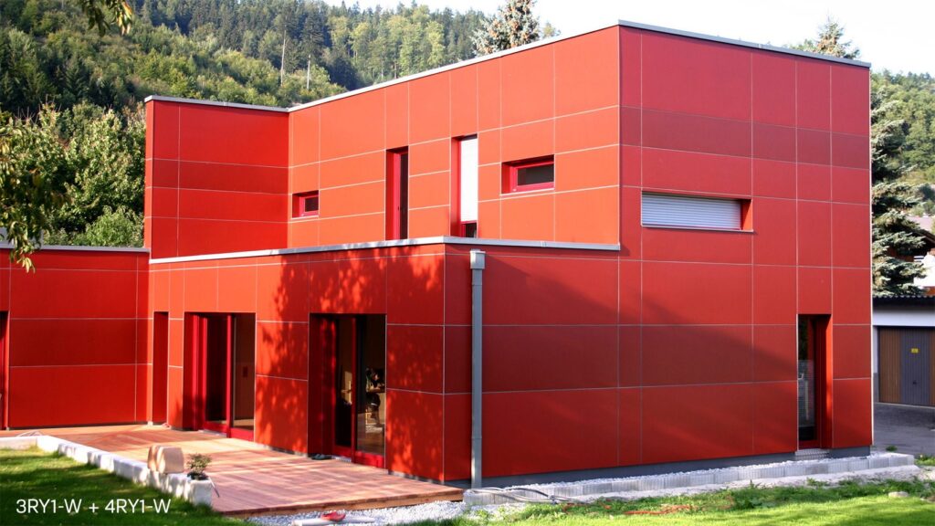 A modern, red cube-shaped house with large windows and a flat roof. It is surrounded by greenery, including trees and a well-maintained lawn. There is a wooden deck in front of the house. The background features a forested hillside.