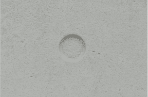 A close-up of a textured, light gray surface featuring a circular indentation in the center. The surface appears to be made of concrete or a similar material with small pores and a slightly rough texture.