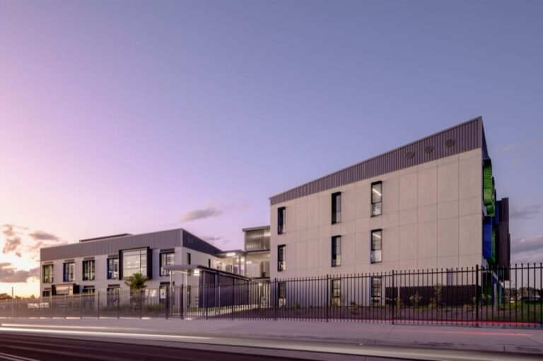 Modern school building at sunset, featuring large windows and a sleek design. A fence runs along the front, with the sky transitioning from purple to pink, suggesting dusk. The architecture is minimalist with clean lines and geometric shapes.